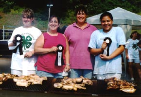 two girls and two women stand behind a grill of smoking chicken
