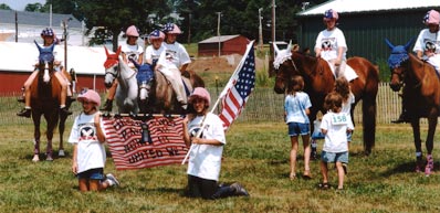 Six children are on horseback while two kneal holding the flag and a red and white striped banner.